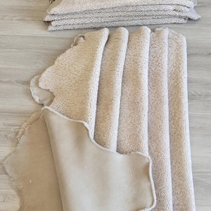 May include: Beige sheepskin rugs, stacked and arranged on a light wood-grain surface. The rugs have a soft, textured surface and a smooth, suede-like underside. The edges of the rugs are irregularly shaped.