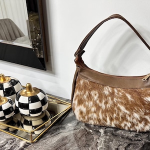 May include: A brown and white cowhide shoulder bag with a brown leather strap and trim. The bag is on a marble surface next to decorative black and white checkered pumpkin-shaped objects with gold stems, displayed on a gold-framed mirrored tray.