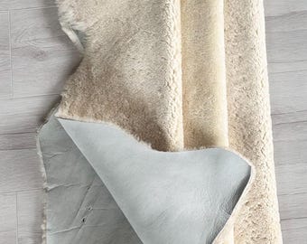 May include: A cream-colored sheepskin rug with a light gray leather backing. The rug is folded over, revealing the smooth leather underside. The wool is thick and fluffy, with a natural, uneven texture. The rug is lying on a light gray wooden floor.