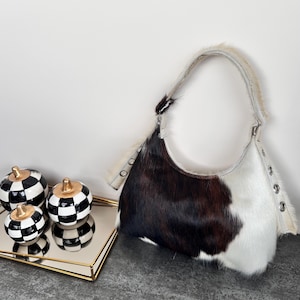May include: A hobo-style shoulder bag crafted from cowhide featuring a distinct color-blocked pattern of dark brown, black, and white fur. The bag includes a single fur-covered strap attached with silver-toned metal grommets on both sides.