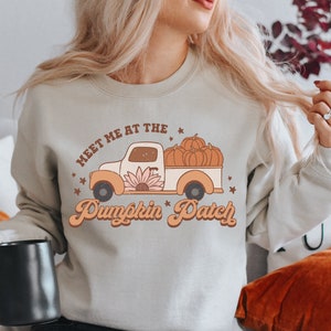 Puede incluir: Una sudadera de color beige claro con un gráfico de estilo vintage de una camioneta que lleva calabazas. El texto en la sudadera dice "Meet me at the Pumpkin Patch".
