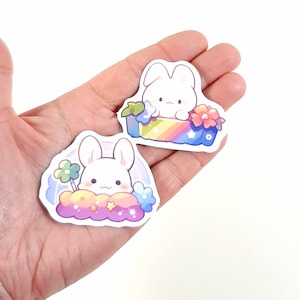 May include: Two stickers featuring a white bunny with pink cheeks. One sticker shows the bunny sitting in a rainbow box with flowers. The other sticker shows the bunny sitting on a cloud with stars and flowers.