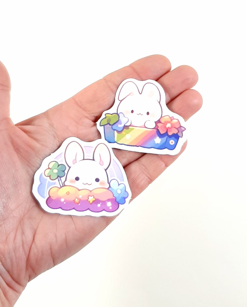 May include: Two stickers featuring a white bunny with pink cheeks. One sticker shows the bunny sitting in a rainbow box with flowers. The other sticker shows the bunny sitting on a cloud with stars and flowers.