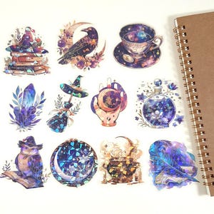 May include: A selection of fantasy-themed stickers, including a raven, teacup, and crystal, in shades of purple, blue, and gold. The stickers have a watercolor effect. A spiral-bound notebook is visible on the right side.