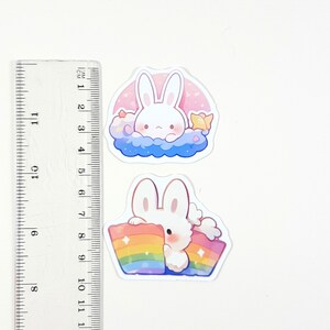 May include: Two stickers featuring a white bunny rabbit. The first sticker shows the bunny in a blue cloud with pink stars and a yellow star. The second sticker shows the bunny holding a rainbow.