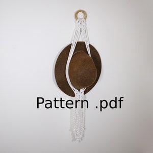 Könnte beinhalten: Ein weißer Makramee-Pflanzenhänger mit einem braunen Hut, der daran hängt. Der Hänger ist an einem Holzring befestigt und hat eine lange Franse. Der Text "Pattern.pdf" ist unter dem Hänger gedruckt.