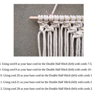 以下が含まれることがあります： 白い紐が結び目模様に結ばれたマクラメの壁掛けのクローズアップ。紐は木製の棒に結ばれています。