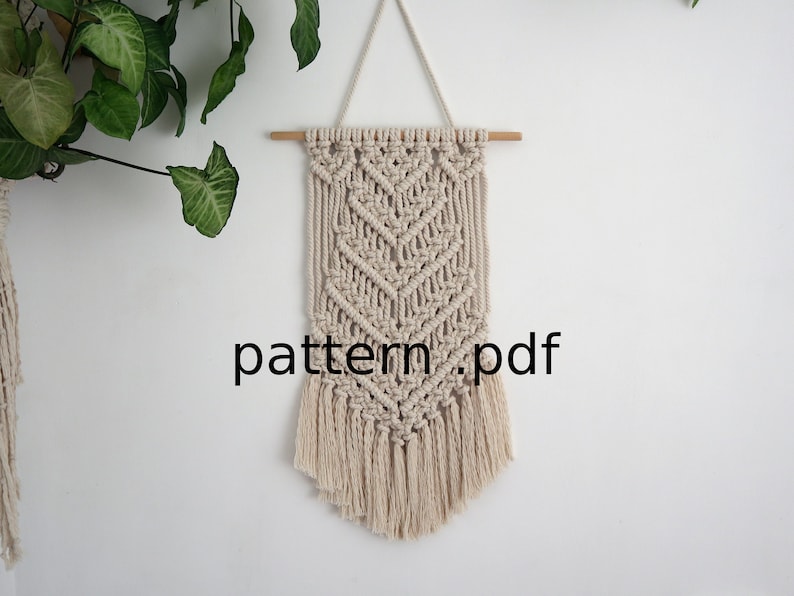 以下が含まれることがあります： 幾何学模様とフリンジが付いたベージュの糸で編まれたマクラメの壁掛け。木製の棒に吊り下げられています。画像には「pattern.pdf」というテキストが表示されています。