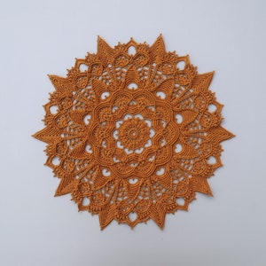 Op de afbeelding: Een groot, rond, gehaakt tafellaken in een warme, gebrande oranje kleur. Het tafellaken heeft een delicaat, ingewikkeld ontwerp met een centraal motief en stralende patronen.