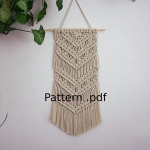 May include: A beige macrame wall hanging with a geometric pattern and fringe. The hanging is attached to a wooden dowel rod. The text "Pattern.pdf" is printed below the hanging.