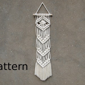 May include: White macrame wall hanging with a geometric pattern and fringe. The hanging is attached to a wooden dowel.