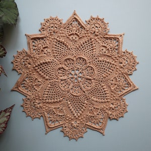 May include: A delicate, peach-colored crocheted doily with a starburst design. The doily has a intricate pattern with a central flower motif.