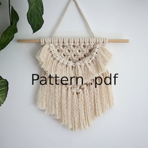 Może przedstawiać: Beżowy wiszący makramowy z geometrycznym wzorem i frędzlami. Wisiorek jest przymocowany do drewnianego pręta. Tekst "Pattern.pdf" jest widoczny na zdjęciu.