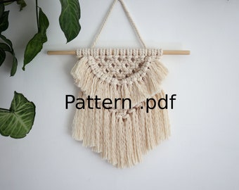 Könnte beinhalten: Ein beigefarbener Makramee-Wandbehang mit geometrischem Muster und Fransen. Der Behang ist an einer Holzleiste befestigt. Der Text "Pattern.pdf" ist auf dem Bild sichtbar.