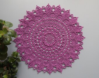 Può includere: Un centrino circolare fatto a mano in una vibrante tonalità di viola. Il lavoro all'uncinetto presenta un intricato design traforato con un bordo smerlato. Il centrino è posto su uno sfondo grigio, con una pianta verde visibile sul lato sinistro.