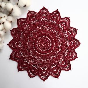 Puede incluir: Un encaje de ganchillo de color rojo oscuro con un delicado patrón floral. El encaje es circular y tiene un centro elevado.
