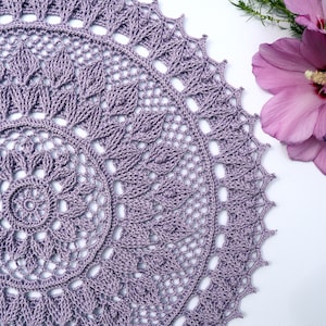 May include: A delicate, crocheted doily in a soft purple hue. The doily features a circular design with intricate details and a central motif.