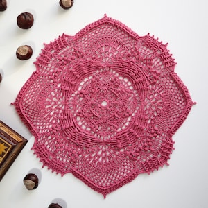 May include: A pink crocheted doily with a delicate floral pattern. The doily is square shaped and has a scalloped edge.
