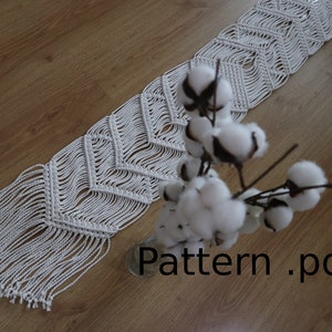 May include: White macrame table runner with a chevron pattern and fringe. The runner is lying on a wooden surface with a vase of cotton flowers.