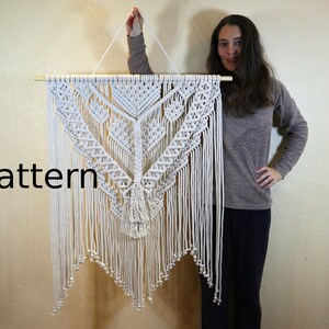 May include: A white macrame wall hanging with a geometric pattern and fringe. The hanging is attached to a wooden dowel rod.