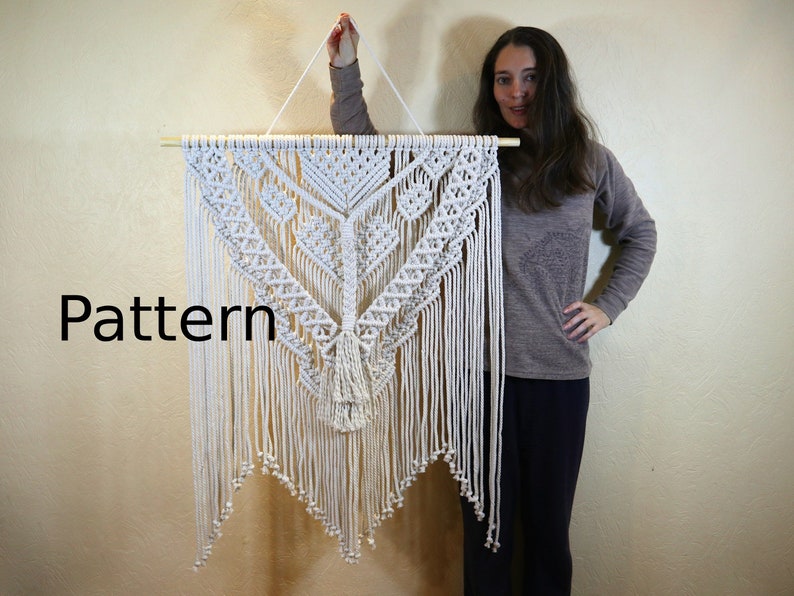 May include: A white macrame wall hanging with a geometric pattern and fringe. The hanging is attached to a wooden dowel rod.