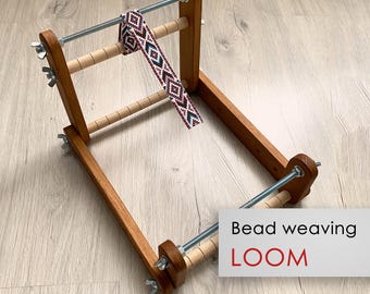 May include: A wooden bead weaving loom with a partially woven beaded strip. The loom has a rectangular frame with adjustable bars and wing nuts. The beaded strip features a geometric pattern in shades of red, black, and white. Text on the image reads "Bead weaving LOOM."