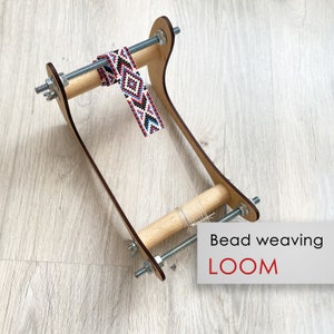 May include: A wooden bead weaving loom featuring two light wood rollers connected by threaded metal rods with silver nuts and wing nuts. A partially woven strip with a geometric pattern in red, white, blue, and black beads is attached.