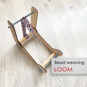 May include: A wooden bead weaving loom featuring two vertical side panels connected by two horizontal wooden dowels and two threaded metal rods with nuts. A partially woven band with a geometric pattern in red, white, black, and blue is attached.