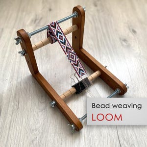 Può includere: Un telaio per perline in legno con aste filettate in metallo e dadi a farfalla. Una striscia di perline parzialmente tessuta con un motivo geometrico in rosso, bianco, nero e blu è fissata ai rulli. Il testo "Bead weaving LOOM" è visibile.