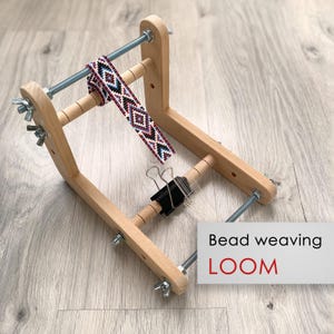 Può includere: Un telaio per perline in legno chiaro con aste filettate in metallo, dadi a farfalla e una clip nera che fissa i fili di ordito bianchi. È attaccata una fascia parzialmente tessuta con un motivo geometrico in rosso, blu, nero e bianco.