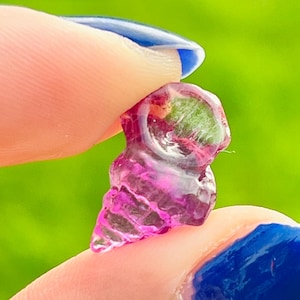 May include: A small, translucent pink and green gemstone is held between two fingers with blue nail polish against a blurred green background. The gemstone has a crystalline structure and a vibrant colour.