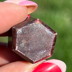 May include: A faceted, dark reddish-brown gemstone with a hexagonal shape. The gem is held between fingers, with a blurred green background. The stone has a slightly iridescent quality, catching the light.