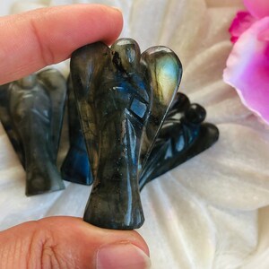Könnte beinhalten: Nahaufnahme mehrerer kleiner, geschnitzter Engelsfiguren. Die Engel sind dunkel, iriserend grau-grün, mit subtilen blauen und goldenen Akzenten. Die Figuren werden in einer Hand gehalten, mit einem weichen, weißen Hintergrund und rosa Blütenblättern.