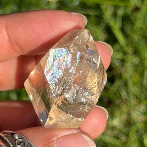 Puede incluir: Un cristal claro y facetado con un tono dorado, sostenido en una mano. La superficie del cristal refleja un arcoíris de colores. El fondo es hierba verde borrosa.
