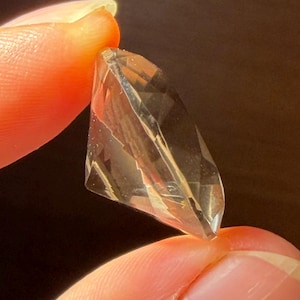 May include: A faceted, transparent, light brown gemstone with a marquise-like shape. The stone features multiple geometric cuts that reflect light, creating internal highlights and shadows throughout its clear, crystalline structure.
