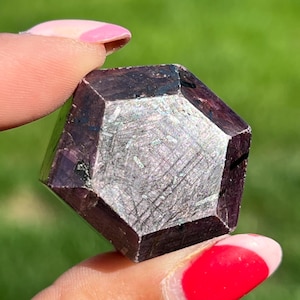May include: A faceted, dark burgundy crystal with a metallic, silver-toned surface. The crystal is held between two fingers against a blurred green background. The crystal's geometric shape is prominent.