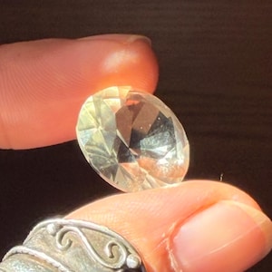 May include: A round, faceted, clear gemstone with a brilliant cut that reflects light. The stone is transparent and colourless, displaying multiple geometric facets across its surface that create a sparkling effect when illuminated from behind.