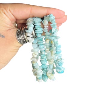 May include: A strand of polished, irregular chip-shaped beads in varying shades of light blue, turquoise, and pale cream. The beads are strung together to form a continuous necklace or bracelet, showcasing a natural, tumbled texture and color.