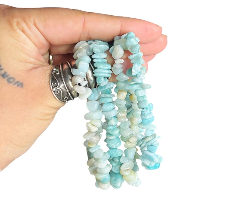 May include: A strand of polished, irregular chip-shaped beads in varying shades of light blue, turquoise, and pale cream. The beads are strung together to form a continuous necklace or bracelet, showcasing a natural, tumbled texture and color.