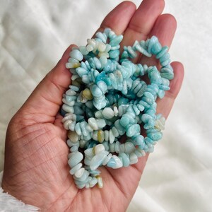 May include: A strand of tumbled, irregular chip beads in various shades of light blue, turquoise, and pale cream. The small, polished stones are strung together, creating a textured, multi-layered appearance with a smooth, glossy finish.