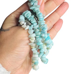May include: A bracelet composed of multiple strands of tumbled, light blue and white stone chips. The stones have a smooth, polished texture and vary in size and shape, creating a layered appearance. The bracelet is secured with a silver-toned metal clasp.