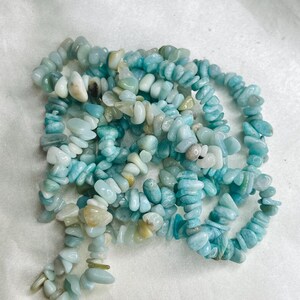 May include: A strand of tumbled, irregular chip beads in shades of light blue, turquoise, and pale green. The polished stones are strung together, creating a textured, multi-layered appearance with variations in opacity and color saturation.