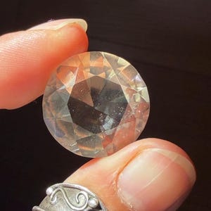 May include: A round, faceted, clear gemstone with a brilliant cut. The stone is transparent and reflects light through its multiple geometric surfaces, showcasing a high level of clarity and a polished finish that creates a sparkling appearance.
