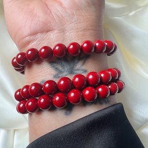 Könnte beinhalten: Zwei rote Perlenarmbänder. Jedes Armband besteht aus mehreren runden, glänzenden Perlen. Die Armbänder werden an einem Handgelenk getragen, mit einem schwarzen Ärmel im Hintergrund. Die Perlen sind tiefrot.