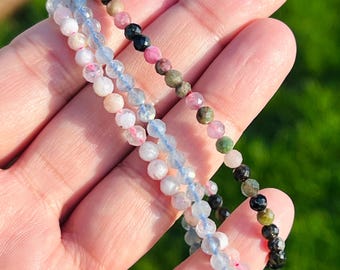 May include: Two delicate beaded bracelets featuring small, faceted, round gemstones. One bracelet consists of translucent light blue and pale pink beads, while the other features a mix of dark green, black, and muted pink faceted stone beads.