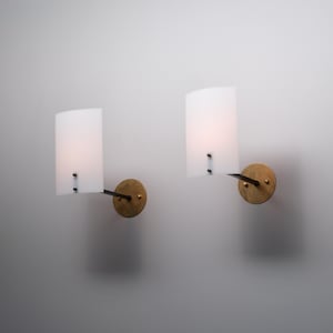 Può includere: Due applique da parete con paralumi rettangolari e curvi in vetro bianco satinato. Ogni paralume è fissato a un sottile braccio in metallo scuro collegato a una piastra circolare in ottone spazzolato. Le lampade emettono una luce soffusa.