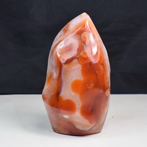 May include: A polished, flame-shaped carnelian stone. The stone displays a mix of orange, red, and white colours, with a smooth, glossy surface. The natural patterns create a unique, decorative piece, ideal for display or collection.