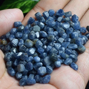 May include: A handful of small, rough-cut blue sapphires. The gemstones vary in shape and size, with a deep blue color and some with hints of gray and brown. The sapphires are unpolished and have a natural, raw appearance.