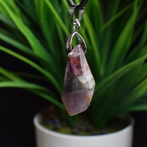 May include: A pendant necklace with a multi-coloured gemstone. The stone has shades of purple, red, white, and grey. It hangs from a silver-toned bail and a black cord. The background is a blurred green plant.