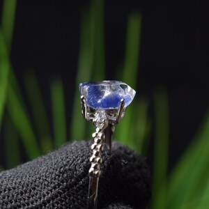 May include: A silver ring with a large, irregular, translucent blue gemstone. The gemstone has a cloudy, textured appearance with darker blue inclusions. The ring's band has a detailed, textured design. The background is blurred.
